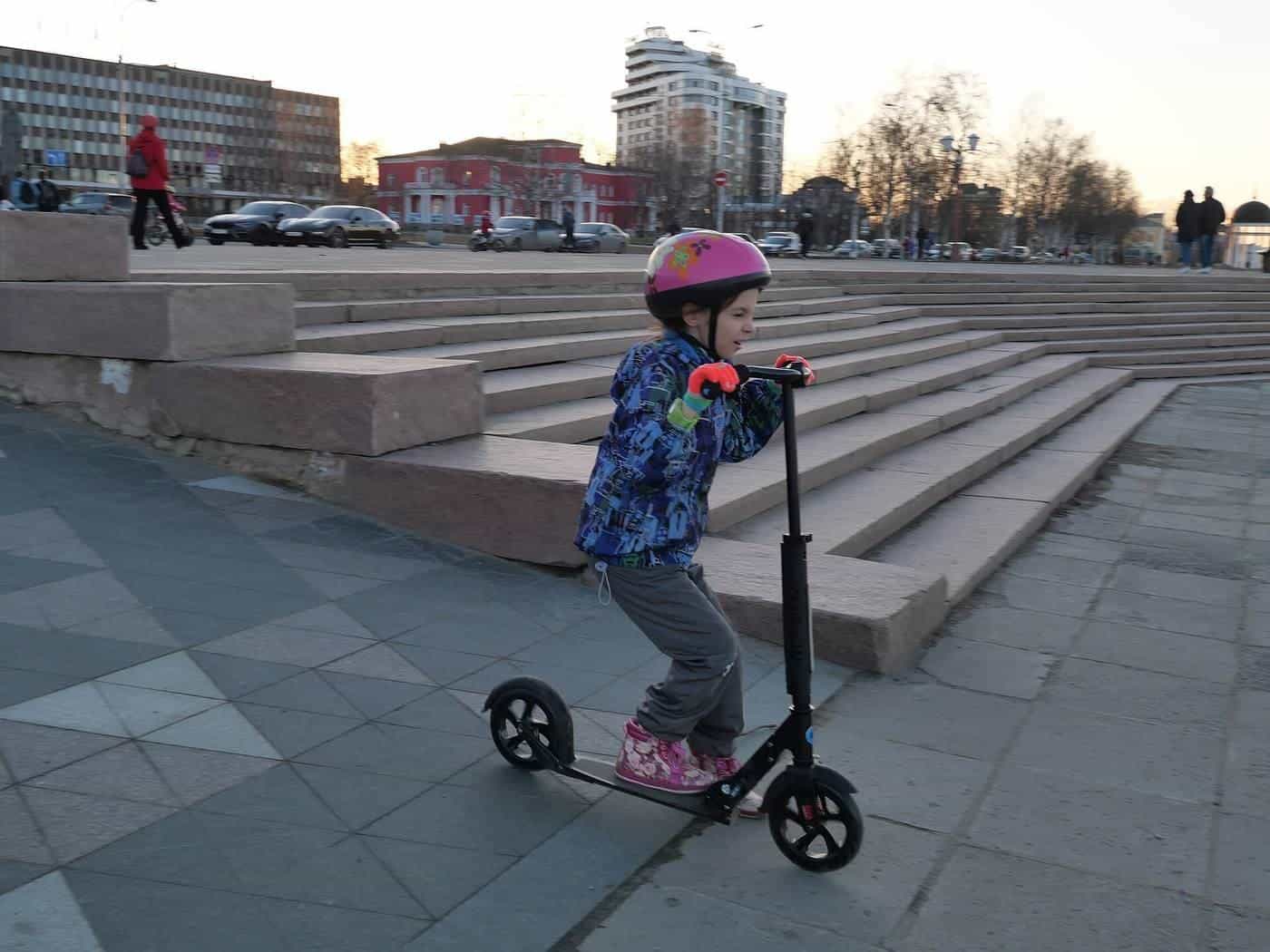 A young child in a pink helmet riding a kick scooter on a paved plaza