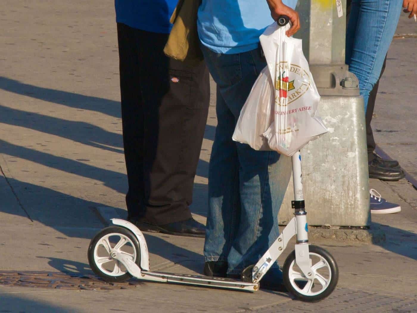 A white folding adult kick scooter held upright on a footpath