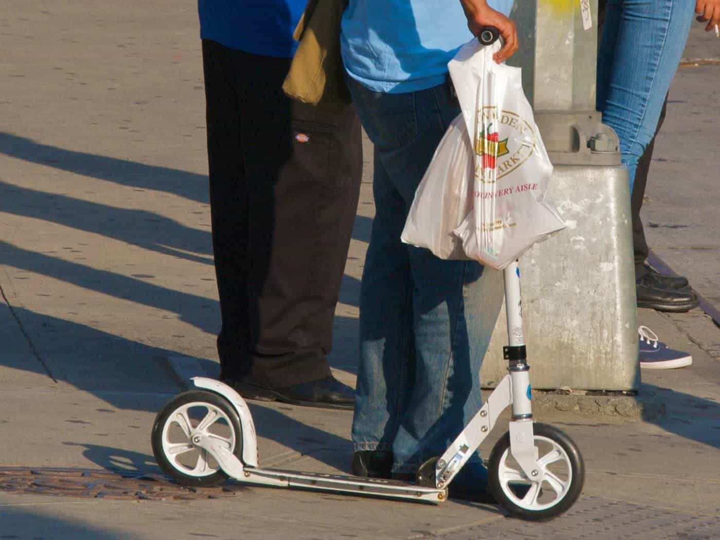 A white folding adult kick scooter held upright on a footpath