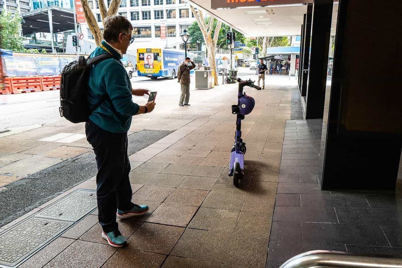 A man holding his phone beside an e-scooter parked on a footpath