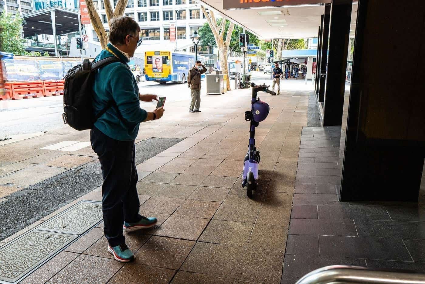 A man holding his phone beside an e-scooter parked on a footpath